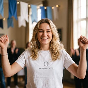 Op de afbeelding: Een wit T-shirt met korte mouwen voorzien van een kleine zwarte lauwerkransafbeelding gecentreerd boven de tekst I'M THE GREATEST in een eenvoudig schreeflettertype. Het shirt heeft een ronde hals en is gemaakt van effen witte stof.