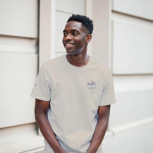 Op de afbeelding: Een lichtgrijs T-shirt met korte mouwen en een kleine afbeelding van een vintage radio op de linkerborst. De radio-illustratie is zwart-wit met een dunne antenne. Een klein logo is zichtbaar op de rechtermouw van het kledingstuk.