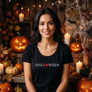 Op de afbeelding: Een zwart T-shirt met korte mouwen en de witte tekst HALLOWEEN op de borst, waarbij een klein rood grafisch element de letter O vervangt. Het shirt heeft een ronde hals en is gemaakt van een effen zwarte stof.