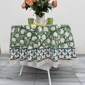 May include: A round tablecloth featuring a green background with white floral patterns, a blue and green scalloped border, and a brown striped edge. The tablecloth is draped over a white table with a vase of peach roses and two green mugs.