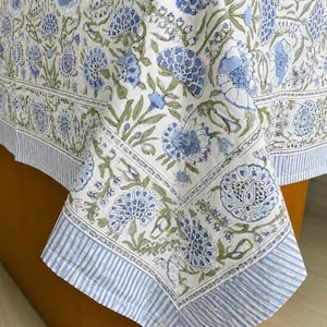 May include: A white tablecloth featuring a repeating floral pattern in shades of blue and sage green. The fabric has a wide border with a blue and white vertical stripe design, framing the central floral motif with intricate botanical details.