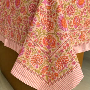 May include: A tablecloth featuring a light peach background with a vibrant floral pattern in shades of orange, hot pink, and green. The fabric includes a decorative border with a repeating striped design in white and light pink along the edges.