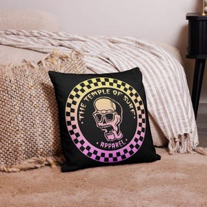 May include: Black square pillow with a circular design featuring a skull wearing sunglasses, with the text "THE TEMPLE OF SURF" and "APPAREL". The design has a gradient of yellow, pink, and purple, with a checkered border.