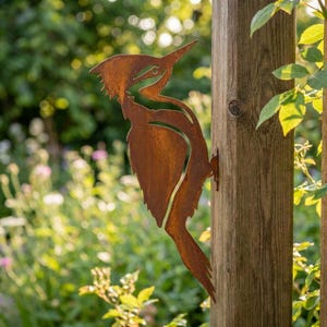 May include: A rusty brown metal woodpecker sculpture attached to a wooden post. The bird is cut from metal and has a detailed design, including a pointed beak and textured feathers. The sculpture is mounted on a wooden post in a garden setting.