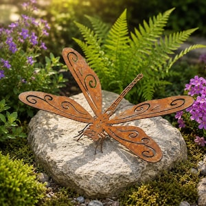 May include: A large, rusty-brown metal dragonfly sculpture with intricate wing designs rests on a gray stone. The dragonfly is surrounded by lush greenery, including ferns and purple flowers, creating a natural garden accent.