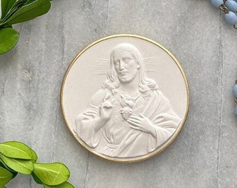 May include: A white ceramic medallion with a raised image of Jesus Christ with his hands over his heart. The medallion is surrounded by a gold trim. A blue rosary with a silver crucifix is also pictured.