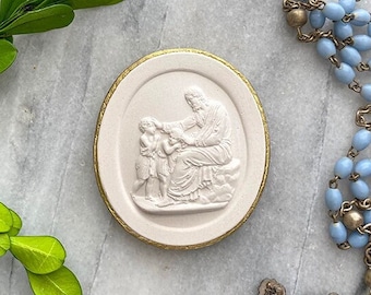 May include: An oval, off-white religious medallion with a gold border, depicting a biblical scene. A light blue rosary and a silver crucifix are also present. Green leaves are arranged around the objects on a marble surface.