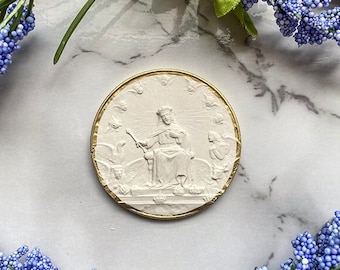 May include: A white ceramic medallion with a gold rim, featuring a raised image of a person sitting on a throne with a crown and halo. The person is surrounded by angels.