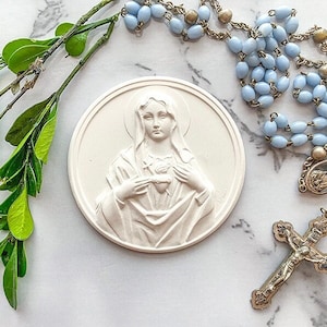 May include: A white ceramic plaque with a relief sculpture of the Virgin Mary holding her heart. The plaque is surrounded by a blue and gold rosary with a crucifix.