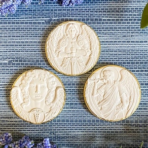 May include: Three cream-colored, round medallions with gold rims. Each medallion features a detailed relief of a figure with wings, including an angel and a person holding a sword. The background includes blue flowers.