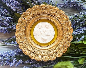 May include: A gold-colored, ornate framed relief sculpture featuring a religious scene. The relief is white, set against a yellow background. The frame is round with intricate detailing. The sculpture is surrounded by purple lavender and green foliage.