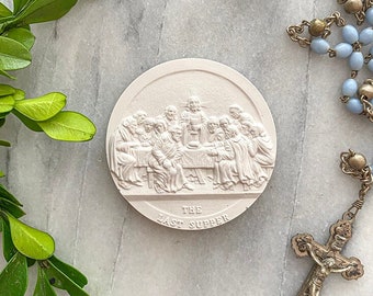 May include: A round, off-white relief sculpture depicting "The Last Supper" with text below. A light blue rosary with a crucifix and green leaves are arranged around the sculpture on a marble surface.
