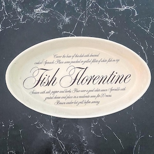 Puede incluir: Una fuente ovalada de cerámica blanquecina con la inscripción "Fish Florentine" en una elegante escritura. La fuente presenta una receta impresa alrededor del borde, con instrucciones para preparar el plato.