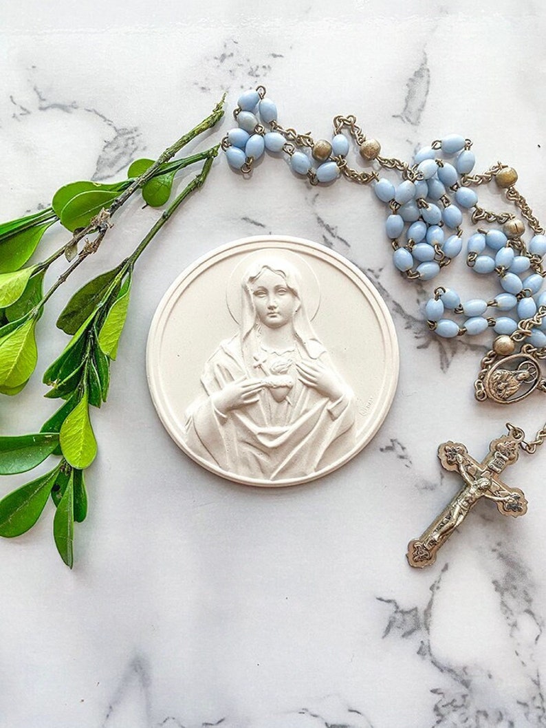 May include: A white ceramic plaque with a relief sculpture of the Virgin Mary holding her heart. The plaque is surrounded by a blue and gold rosary with a crucifix.