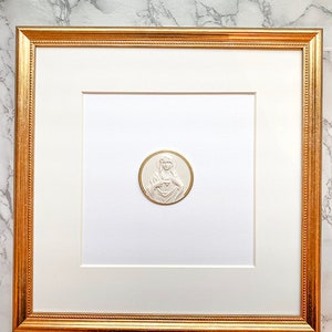 May include: A white framed picture with a gold frame. The picture is of a white, embossed image of a woman with her hands clasped over her heart. The image is on a white background.