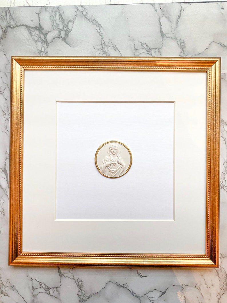 May include: A white framed picture with a gold frame. The picture is of a white, embossed image of a woman with her hands clasped over her heart. The image is on a white background.