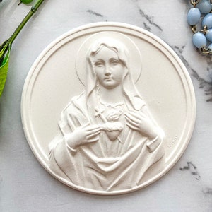 May include: White plaster plaque with a raised image of the Virgin Mary holding a heart with a cross. The plaque is round and has a slightly textured surface.