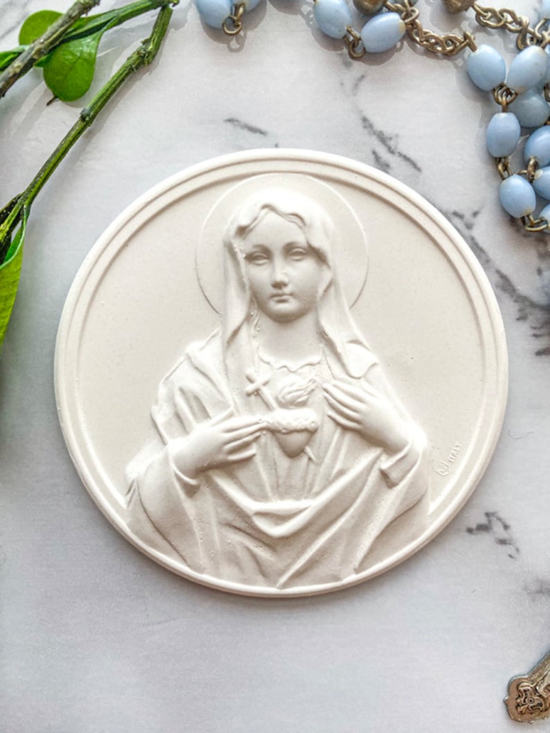 May include: White plaster plaque with a raised image of the Virgin Mary holding a heart with a cross. The plaque is round and has a slightly textured surface.