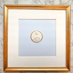 May include: A white framed picture with a gold trim. The picture contains a white, round, embossed image of the Virgin Mary with a heart on her chest.