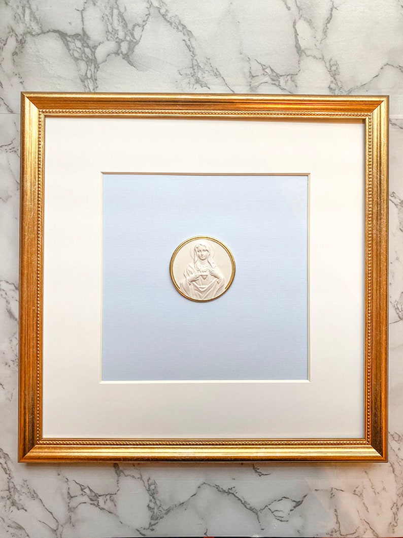 May include: A white framed picture with a gold trim. The picture contains a white, round, embossed image of the Virgin Mary with a heart on her chest.
