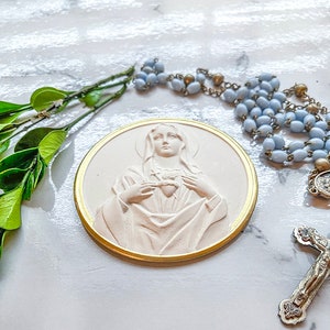 May include: A white plaster relief of the Virgin Mary with a gold trim. She is holding her heart in her hands. The relief is on a white surface with a blue rosary and a silver cross.