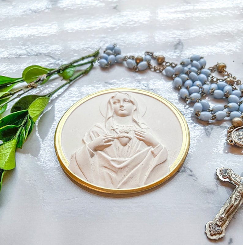 May include: A white plaster relief of the Virgin Mary with a gold trim. She is holding her heart in her hands. The relief is on a white surface with a blue rosary and a silver cross.