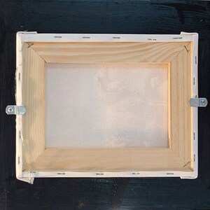 Puede incluir: La parte posterior de un cuadro de lienzo enmarcado con un marco de madera y un lienzo blanco. El marco tiene dos ganchos met&aacute;licos para colgar en los lados.