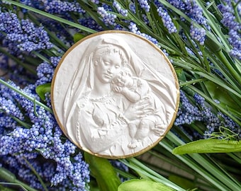 Könnte beinhalten: Weißer Gipsrelief der Jungfrau Maria, die ein Jesuskind hält. Das Relief ist in einen goldenen Kreis gefasst und von blauen und grünen Blumen umgeben.