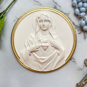 May include: A white plaster relief of the Virgin Mary with a gold-colored frame. She is wearing a white robe and holding a heart in her hands. The heart is pierced with a sword.