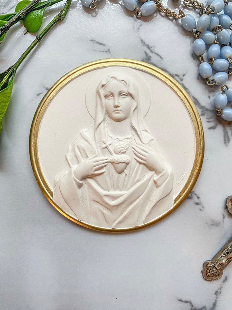 May include: A white plaster relief of the Virgin Mary with a gold-colored frame. She is wearing a white robe and holding a heart in her hands. The heart is pierced with a sword.