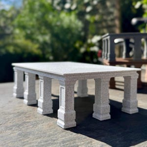 May include: A rectangular, light-colored stone table with a speckled surface. The table is supported by six rectangular columns with decorative detailing. The table is outdoors, with a blurred green background.