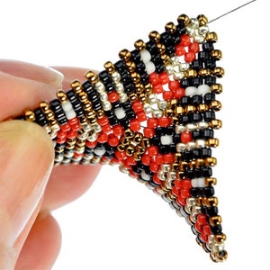 May include: A close-up of a beaded triangle pendant with a black, white, red, gold, and silver color scheme. The pendant is being held by a hand.