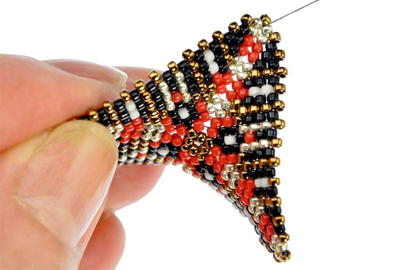May include: A close-up of a beaded triangle pendant with a black, white, red, gold, and silver color scheme. The pendant is being held by a hand.