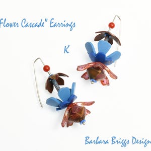 Op de afbeelding: Handgemaakte oorbellen met een bloemendesign. De oorbellen hebben blauwe, bruine en rode bloemvormige elementen, met een kleine rode kraal en een zilveren haak. De tekst "Flower Cascade" Earrings is zichtbaar.