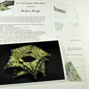 May include: A close-up of a green and gold beaded wrist wrap, designed as a "Sea Dragon" by Barbara Briggs. The wrist wrap is made with Delica beads and features a faux seashell accent.