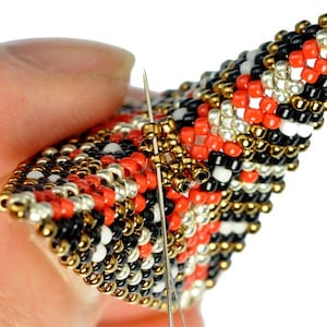 May include: Close-up of a beaded project with a needle and thread going through the beads. The beads are red, black, white, gold, and silver.