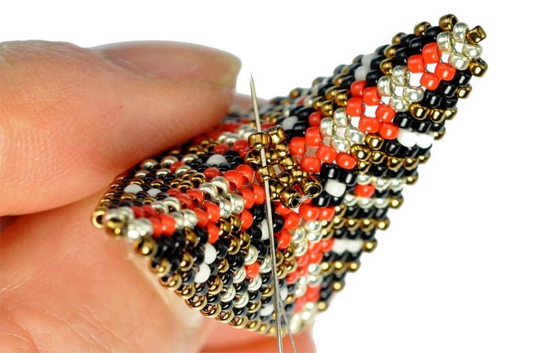 May include: Close-up of a beaded project with a needle and thread going through the beads. The beads are red, black, white, gold, and silver.