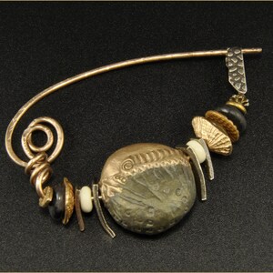 May include: A gold and silver brooch with a large round pendant featuring a textured design. The brooch is adorned with smaller beads and charms in black, gold, and white.