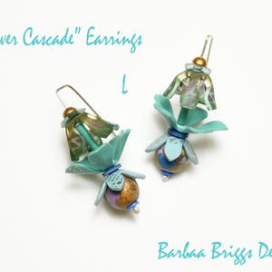Op de afbeelding: Een paar handgemaakte oorbellen met een bloemenontwerp. De oorbellen hebben teal, goud en paarse accenten, met de tekst "Flower Cascade" Earrings en "Barbaa Briggs Designs". De oorbellen hebben een unieke, artistieke stijl.
