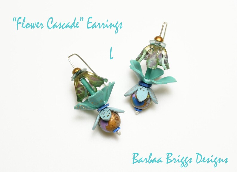 Op de afbeelding: Een paar handgemaakte oorbellen met een bloemenontwerp. De oorbellen hebben teal, goud en paarse accenten, met de tekst "Flower Cascade" Earrings en "Barbaa Briggs Designs". De oorbellen hebben een unieke, artistieke stijl.