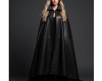 Op de afbeelding: Een lange zwarte leren cape met een brede kraag en een knoopsluiting. De cape wordt gedragen over een zwarte jurk, wat een dramatische en elegante look creëert. Het model heeft lang blond haar en staat tegen een donkergrijze achtergrond.