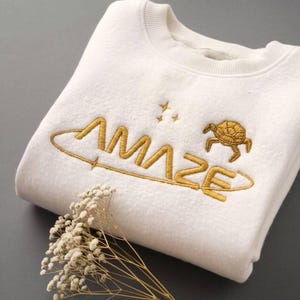 Peut inclure: Un sweat-shirt blanc plié, orné de broderies dorées. Le mot "AMAZE" est brodé en lettres dorées, avec une tortue et des étoiles. Le design est entouré d'une ligne dorée.