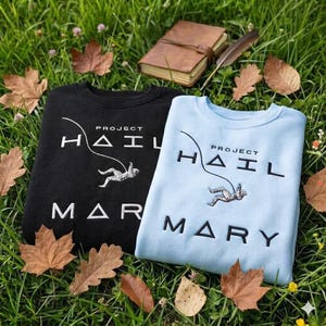 Peut inclure: Deux sweatshirts, un noir et un bleu clair, avec le texte "PROJECT HAIL MARY" en blanc. Chaque sweat-shirt présente un graphisme d'une personne suspendue par une ligne. Ils sont posés sur de l'herbe avec des feuilles mortes.