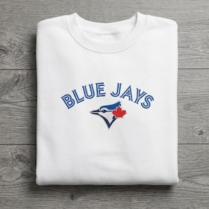 May include: White sweatshirt featuring the text "BLUE JAYS" in blue, arched lettering. Below the text is a blue bird logo with a red maple leaf. The sweatshirt is made of a soft material.