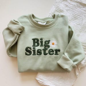 May include: A light green sweatshirt with the embroidered text "Big Sister" in dark green, accompanied by a daisy graphic. The sweatshirt has a crew neck and ribbed cuffs.