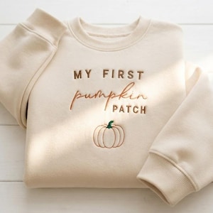 May include: A beige crewneck sweatshirt featuring the embroidered text "MY FIRST pumpkin PATCH" in brown and orange thread, accompanied by a small embroidered pumpkin graphic with a green stem centered below the lettering.
