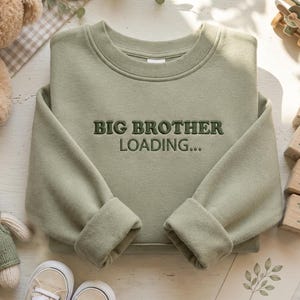 May include: A sage green long-sleeved sweatshirt featuring the embroidered text "BIG BROTHER LOADING..." in a darker green font. The garment has a crew neckline and cuffed sleeves, presented as a flat lay item with a neutral, solid-coloured fabric.