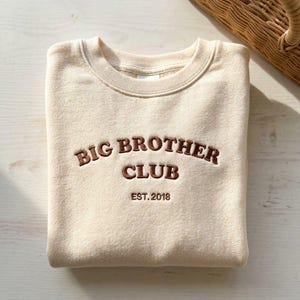 Könnte beinhalten: Beiges Sweatshirt mit den Worten "BIG BROTHER CLUB" in brauner Schrift gestickt. Das Jahr "EST. 2018" ist darunter gestickt. Das Sweatshirt liegt gefaltet auf einer hellen Holzoberfläche, im Hintergrund ein Weidenkorb.