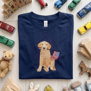 Op de afbeelding: Een marineblauw t-shirt met een borduursel van een golden retriever die een kleine Amerikaanse vlag in zijn bek houdt. Het t-shirt is omringd door diverse kinderspeeltjes, waaronder houten blokken, speelgoedauto's, een teddybeer en een pluche konijn.