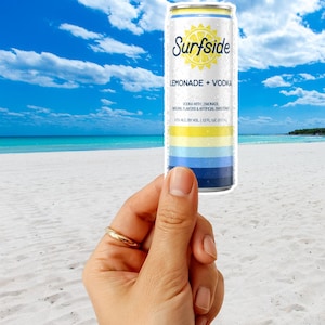 May include: A can of Surfside Lemonade + Vodka held against a beach backdrop. The can features a white top with the brand name and a yellow sun graphic. Below are horizontal stripes of yellow, blue, and navy. The can contains 12 fl oz.